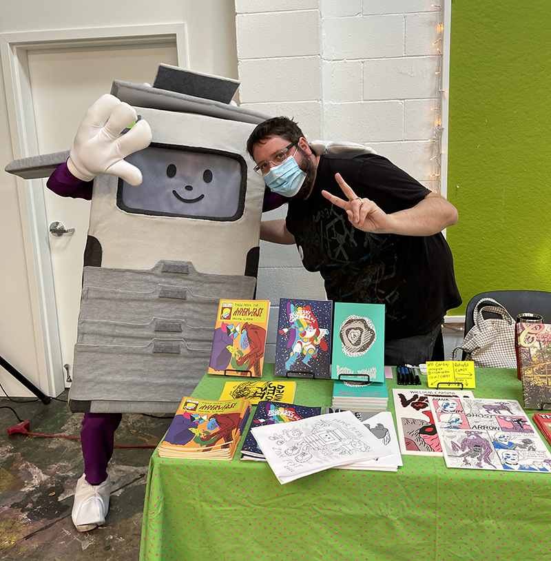 Photo of Will Cardini at Paper Plains Zine Fest, standing behind his table of comics, hugging Jammy the mascot, a person in a full-body cartoonist risograph costume