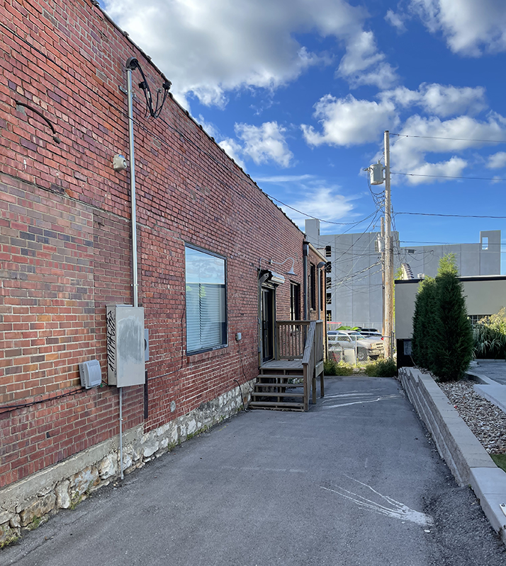 Photo of an alley with a brick wall on the left and blue sky with a few clouds above. There's a wooden staircase towards the end.